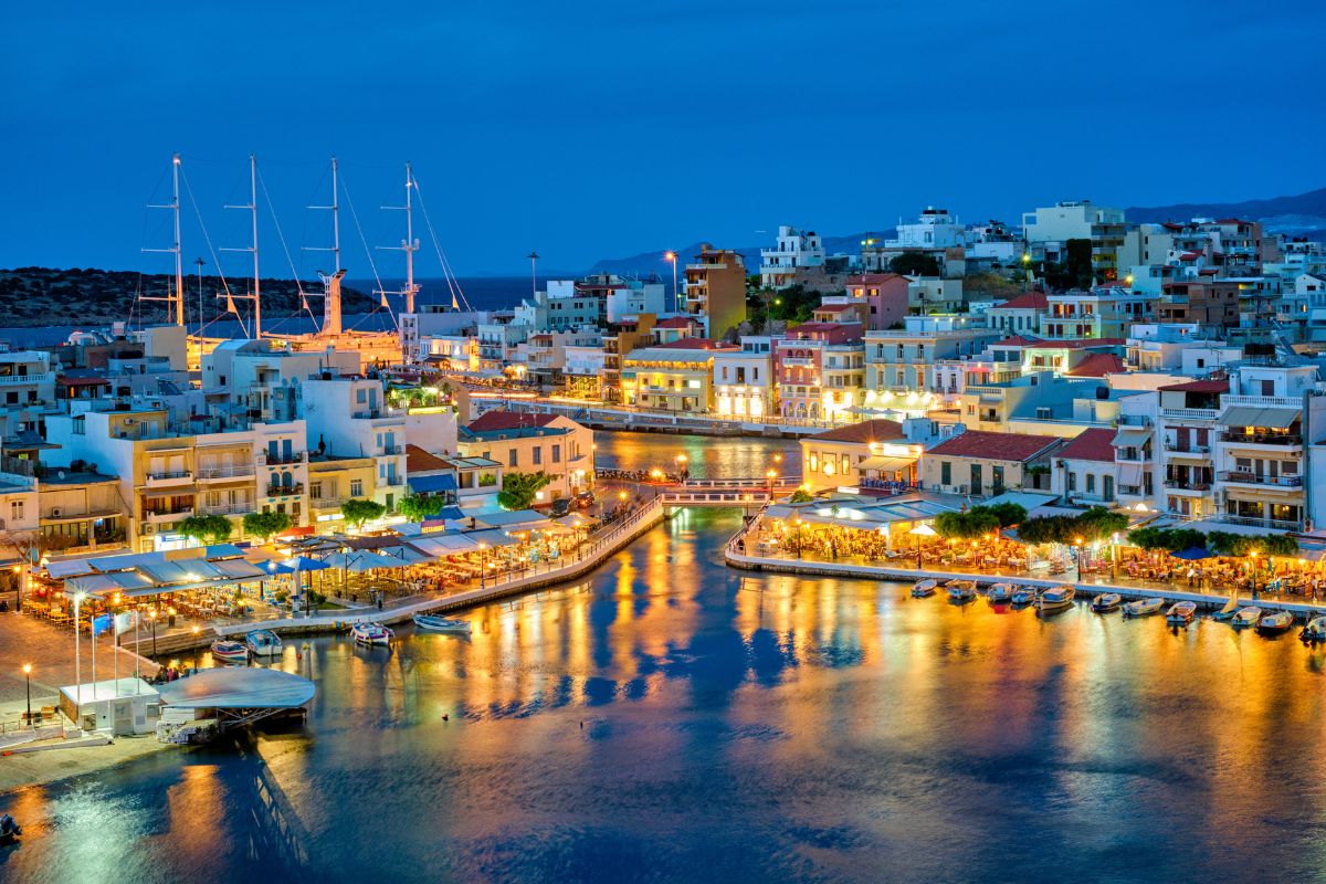 A vibrant evening view of Agios Nikolaos in Crete, showcasing illuminated buildings, boats docked along the waterfront, and a tranquil harbor reflecting the lights.