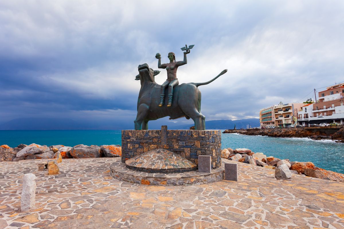 A statue depicting a figure riding a bull stands on a stone platform by the sea in Agios Nikolaos, Crete, with buildings and a cloudy sky in the background.