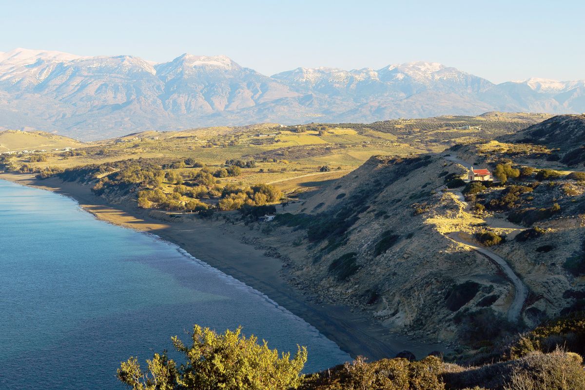 A serene coastal landscape in Crete during winter, with a calm sea gently meeting a sandy beach. The foreground features a hilly terrain with a small house nestled on a slope. In the background, snow-capped mountains rise under a clear sky.