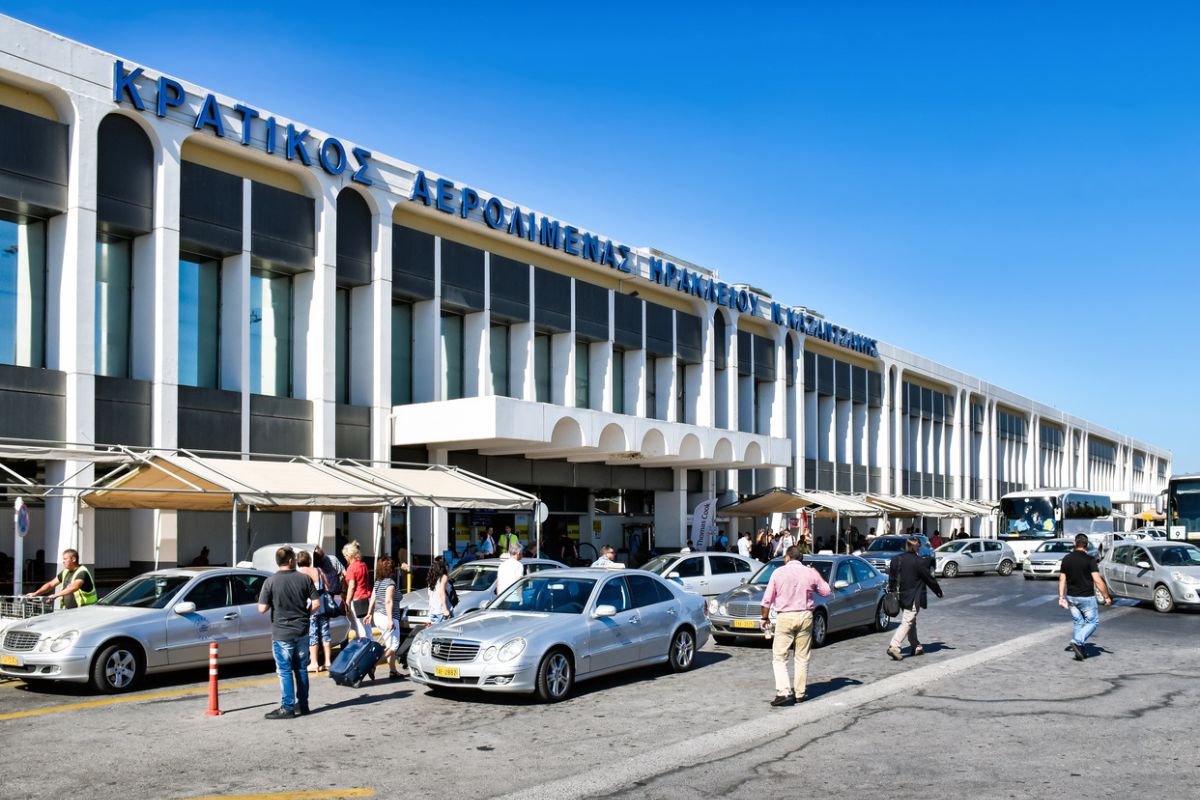 The exterior of Heraklion Airport is shown, with people and vehicles in front of the main terminal building on a clear day. The building features large windows and Greek signage above the entrance_Crete Airport Guide