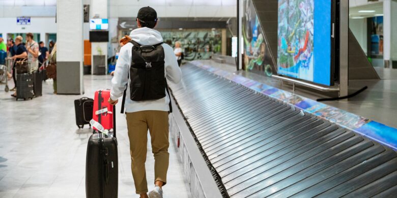 A traveler collects luggage from a baggage carousel at an airport, with several other passengers in the background doing the same. The atmosphere is typical of an airport terminal, with a mix of activity as people retrieve their belongings.