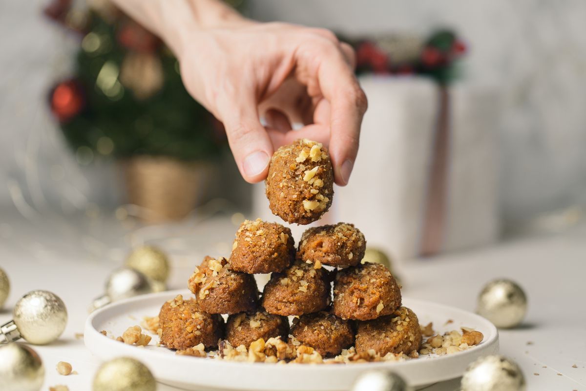 A hand is picking up a melomakarono, a traditional Greek honey cookie, from a pyramid-shaped stack on a plate. The cookies are topped with crushed nuts, and the background includes blurred holiday decorations.