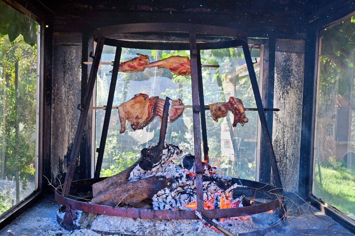 The image shows meat being cooked using the traditional Cretan method called "antikristo," where large pieces of meat are skewered and positioned around an open fire. The setup involves the meat slowly roasting over the flames, creating a rustic and authentic cooking scene. The background includes natural surroundings, adding to the traditional ambiance of the cooking process.