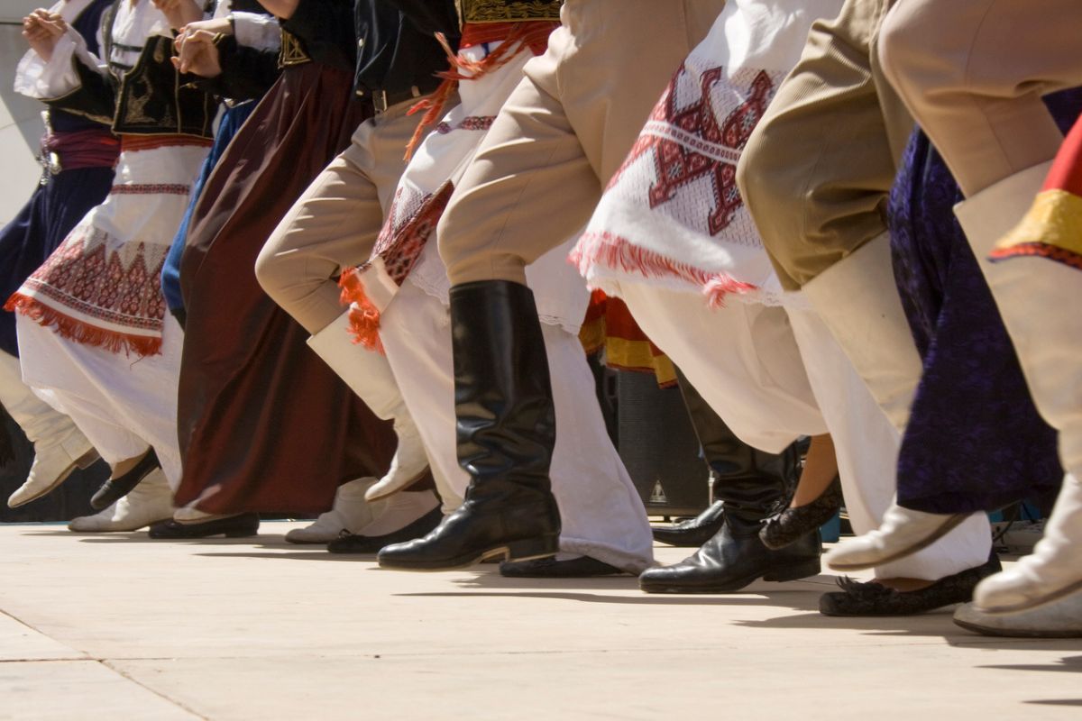 The image shows a group of people participating in a traditional Cretan dance, wearing traditional attire that includes boots, embroidered skirts, and sashes. Their legs are raised in unison, highlighting the rhythmic movement of the dance. The focus is on the lower part of their bodies, emphasizing the coordinated steps and attire typical of Cretan folk culture.