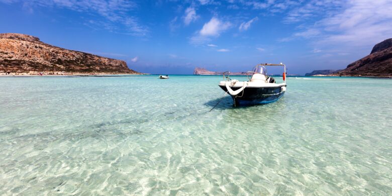 The image shows a serene coastal scene in Crete, with a boat anchored in crystal-clear, shallow turquoise waters. Rugged hills frame the background under a bright blue sky, capturing the tranquil beauty of the island. The overall setting is peaceful and inviting, highlighting Crete's stunning natural landscapes.