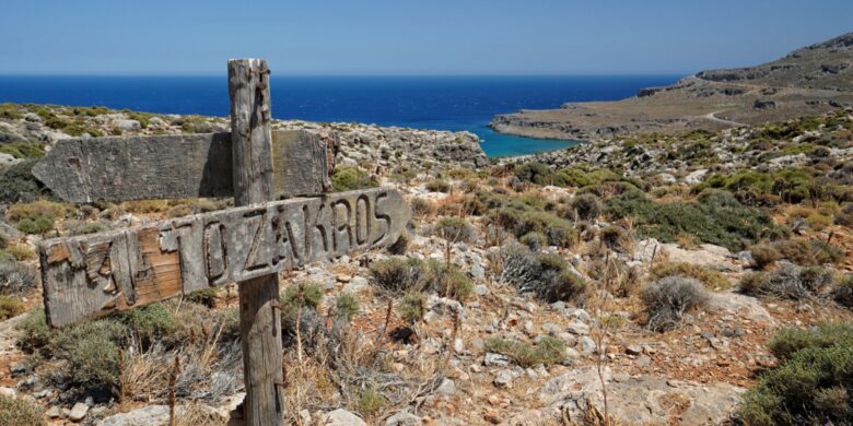 A wooden signpost in a rocky, dry landscape points toward the sea with the word "Zakros" carved into it. In the background, rugged hills meet the coastline, with the blue ocean stretching into the distance under a clear sky.