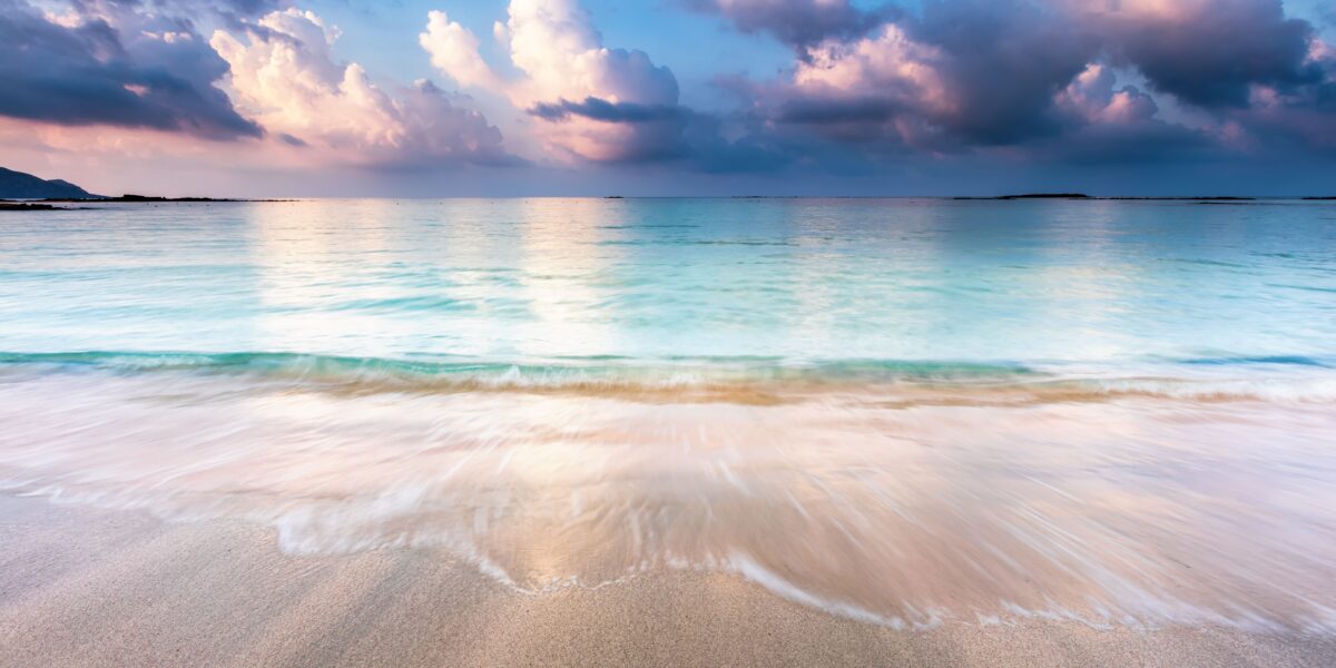 A tranquil shoreline on the island of Crete, with soft waves gently washing onto the sandy beach. The sky is painted in hues of pink and blue, reflecting on the clear, calm waters. The peaceful atmosphere hints at a serene moment just before sunset.