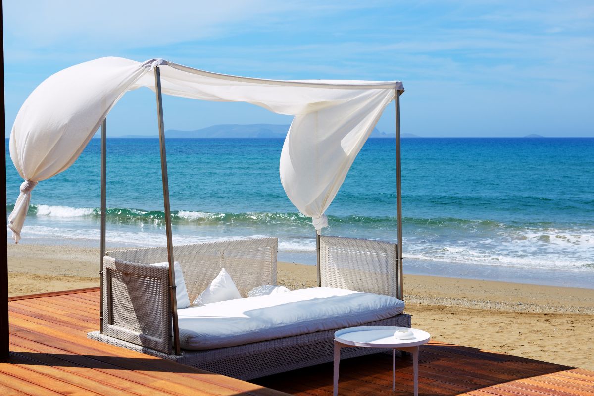 A luxurious canopy daybed overlooks a serene beach with clear blue waters at a hotel in Crete, offering a peaceful seaside retreat.