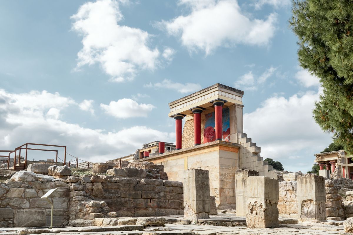 The ancient ruins of Knossos, featuring a restored section with red columns and vibrant frescoes. The remnants of stone walls surround the structure under a bright, cloudy sky.