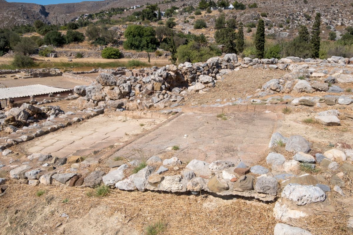 The image shows the ruins of the Minoan Palace of Zakros, with low stone walls and remnants of the palace’s ancient foundations. The surrounding landscape is dry and rocky, with scattered trees and hills in the background under a clear sky.