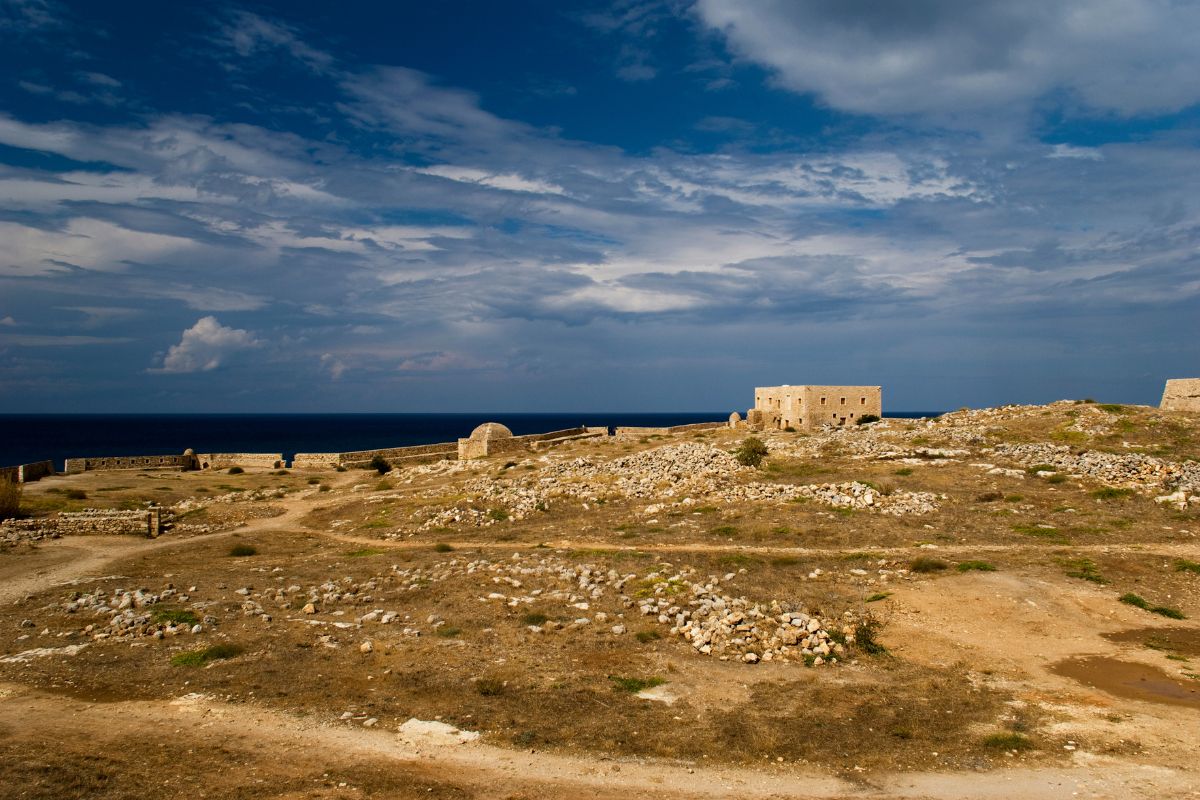 A rugged landscape near Rethymnon with scattered ruins and a stone building, set against a backdrop of deep blue sea and dramatic skies. The dry terrain is dotted with stones, and winding paths stretch across the scene.