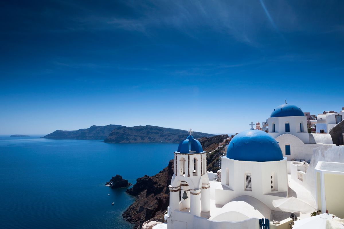 A serene view of Santorini's iconic blue-domed churches against the backdrop of the deep blue Aegean Sea and distant caldera cliffs under a clear sky.