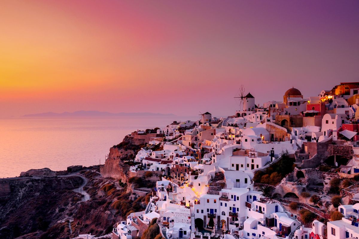 A stunning sunset over Santorini, with the iconic whitewashed buildings and windmills of Oia glowing against the soft hues of the sky and the calm sea in the background.