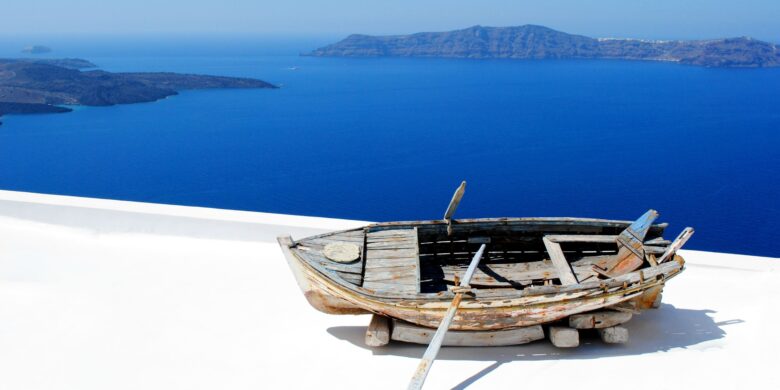 A rustic wooden boat rests on a whitewashed rooftop in Santorini, overlooking the vast, calm Aegean Sea and distant volcanic islands under a bright, clear sky.