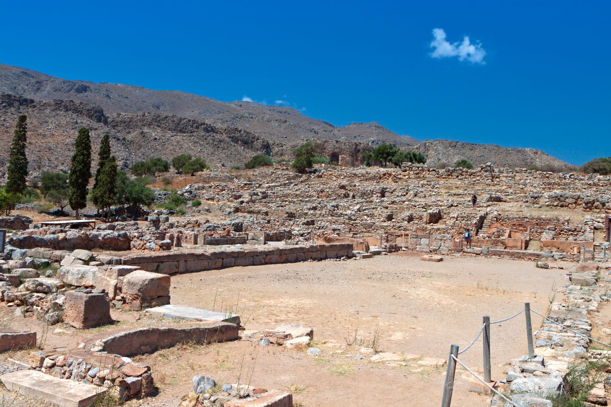 The image shows the Zakros archaeological site, featuring the ruins of ancient stone structures scattered across a dry, rocky landscape. In the background, rugged hills rise under a clear blue sky.