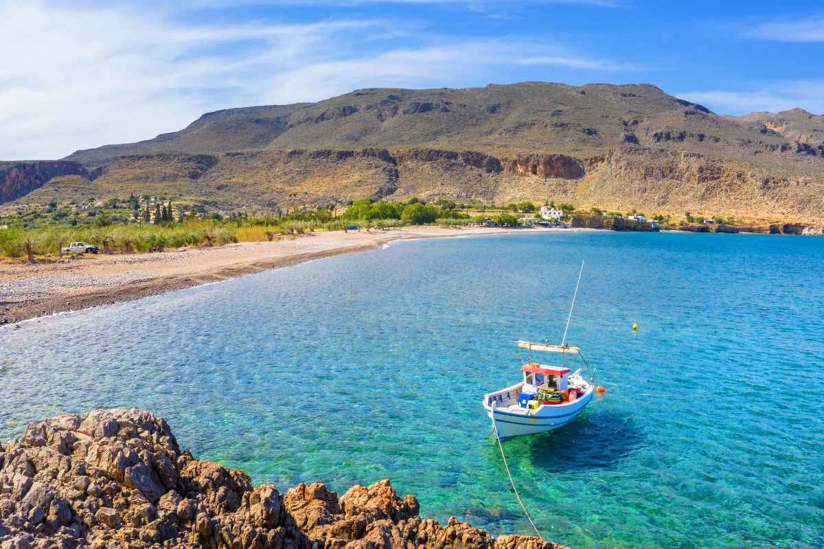 The image shows Zakros Beach with a small boat anchored in clear blue waters near rocky outcrops. The shoreline is sandy and pebbly, backed by sparse greenery and rugged hills under a blue sky.