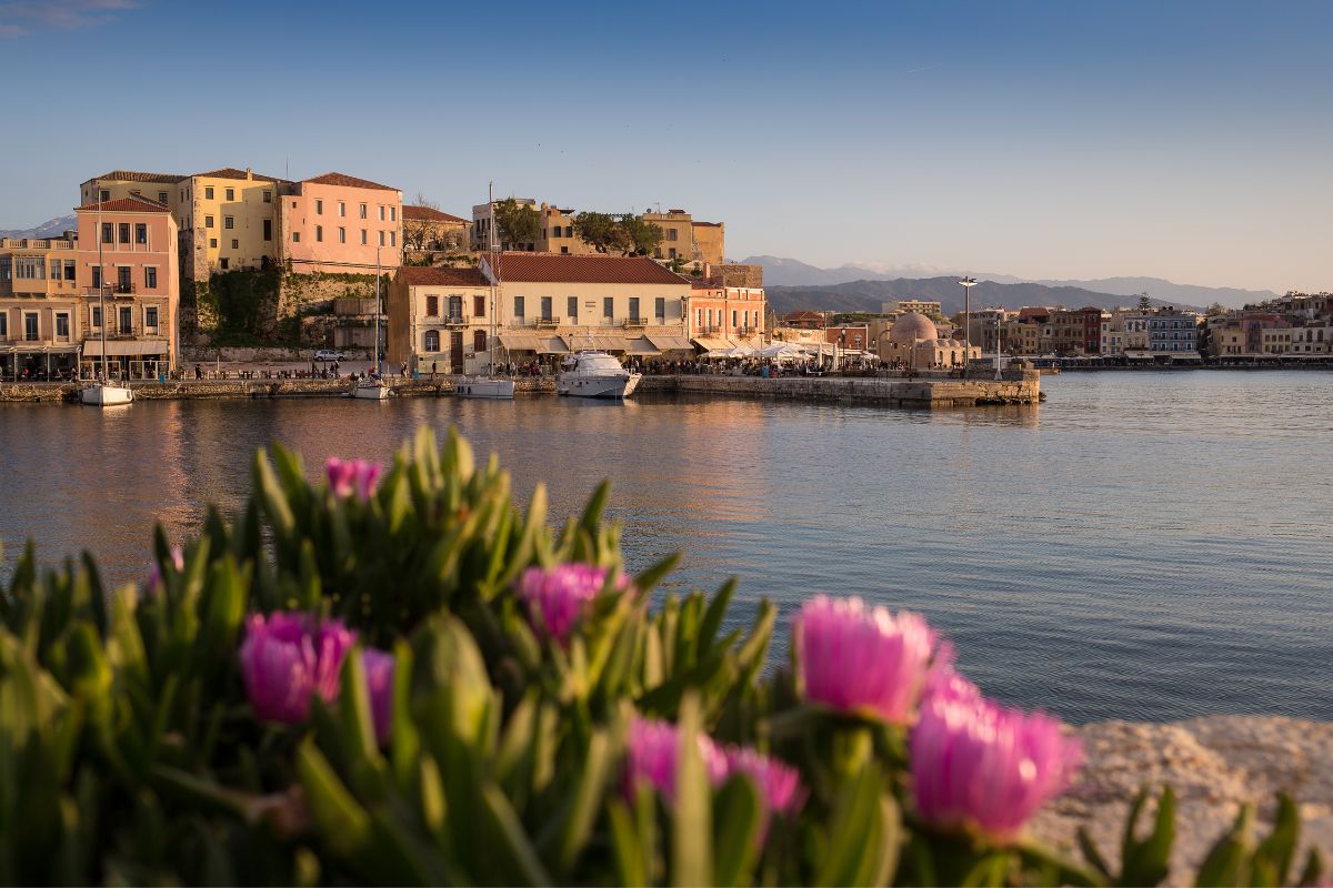 The image shows a peaceful coastal scene in Crete, with pastel-colored buildings lining the waterfront. In the foreground, pink flowers add a vibrant touch, while boats are docked along the harbor. Mountains can be seen in the distance under a clear sky.