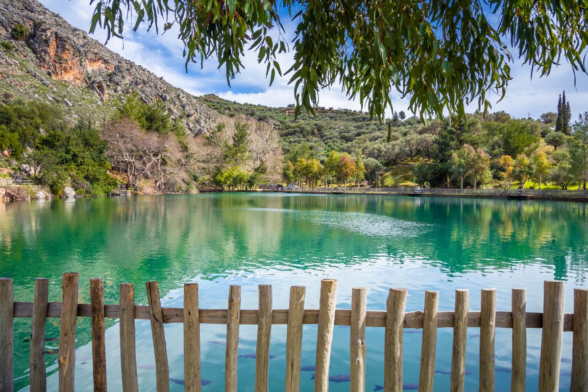 The image shows a serene natural scene in Crete, featuring a calm turquoise lake surrounded by lush greenery and rocky hills. A wooden fence lines the edge of the lake, and trees gently frame the top of the image under a partly cloudy sky.