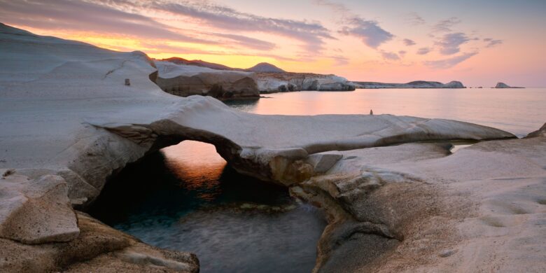 A serene sunset view on Milos Island, showcasing its unique white rock formations and calm waters, with soft orange and pink hues reflecting off the sea.