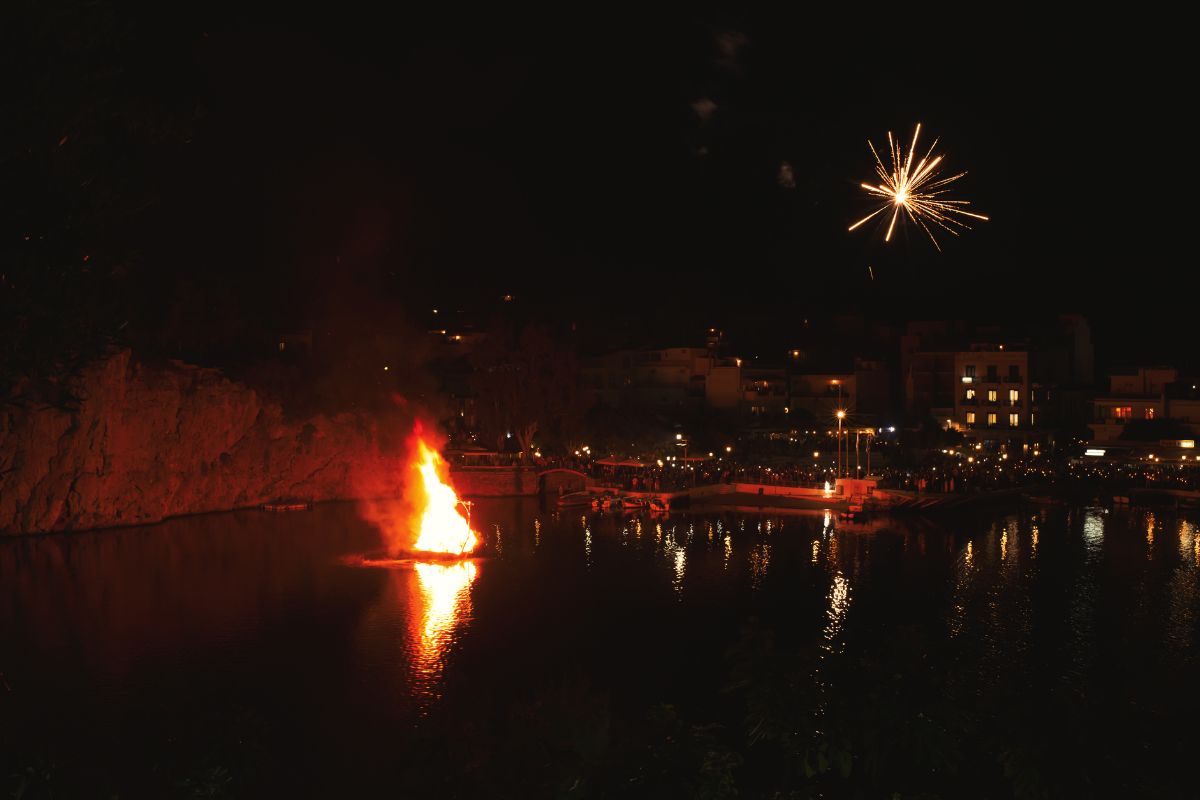 The image depicts a nighttime scene at Agios Nikolaos in Crete, where a large bonfire is burning in the middle of the water, representing the burning of Judas, a traditional Easter celebration. Fireworks light up the sky, and the glow from the flames reflects off the water, while crowds of people watch from the shore.