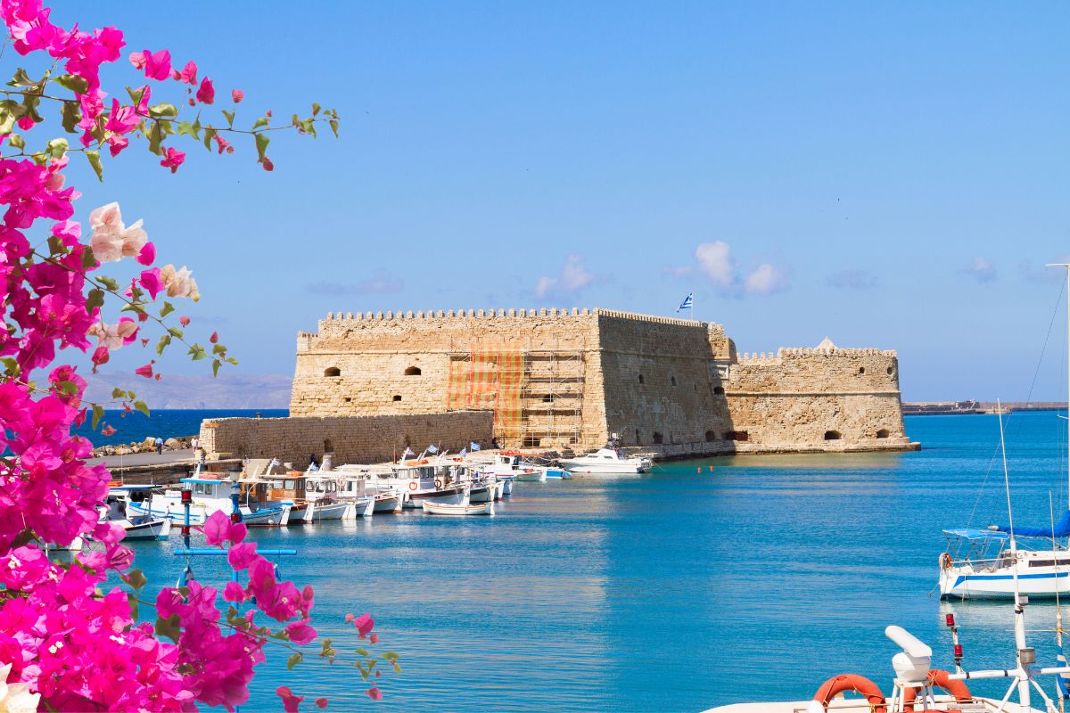 The image shows a bright, sunny view of the Venetian Fortress in Heraklion, Crete, with vibrant pink flowers in the foreground. Boats are docked along the calm blue waters of the harbor, and the clear sky adds to the serene coastal atmosphere.