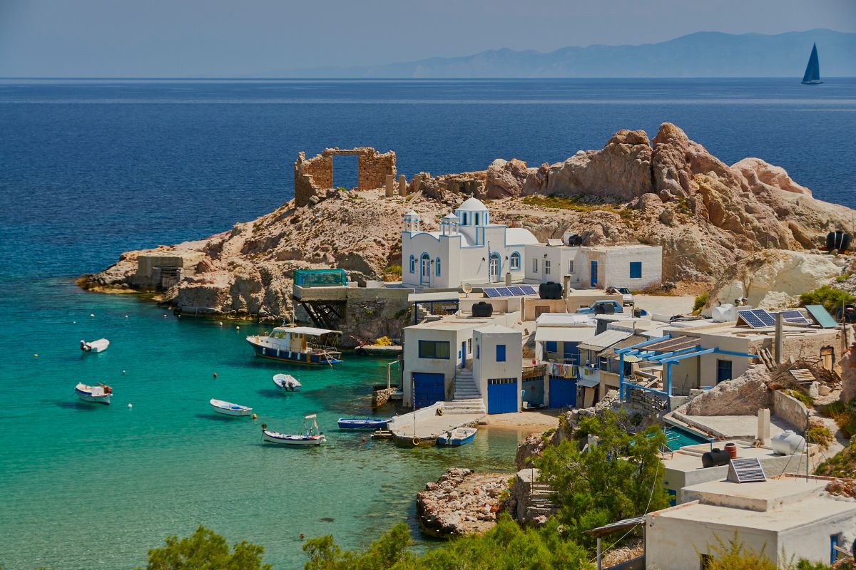 A serene coastal scene on Milos Island, showcasing traditional whitewashed buildings with blue accents near the clear turquoise waters, small fishing boats floating nearby, and a rocky landscape in the background.