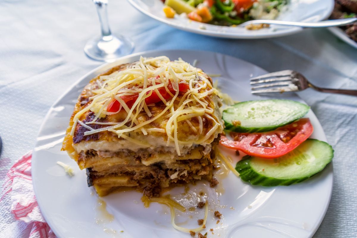 A plate of traditional Greek moussaka topped with shredded cheese and tomato, served with slices of cucumber and tomato on the side. A fork and a glass of water are visible in the background.