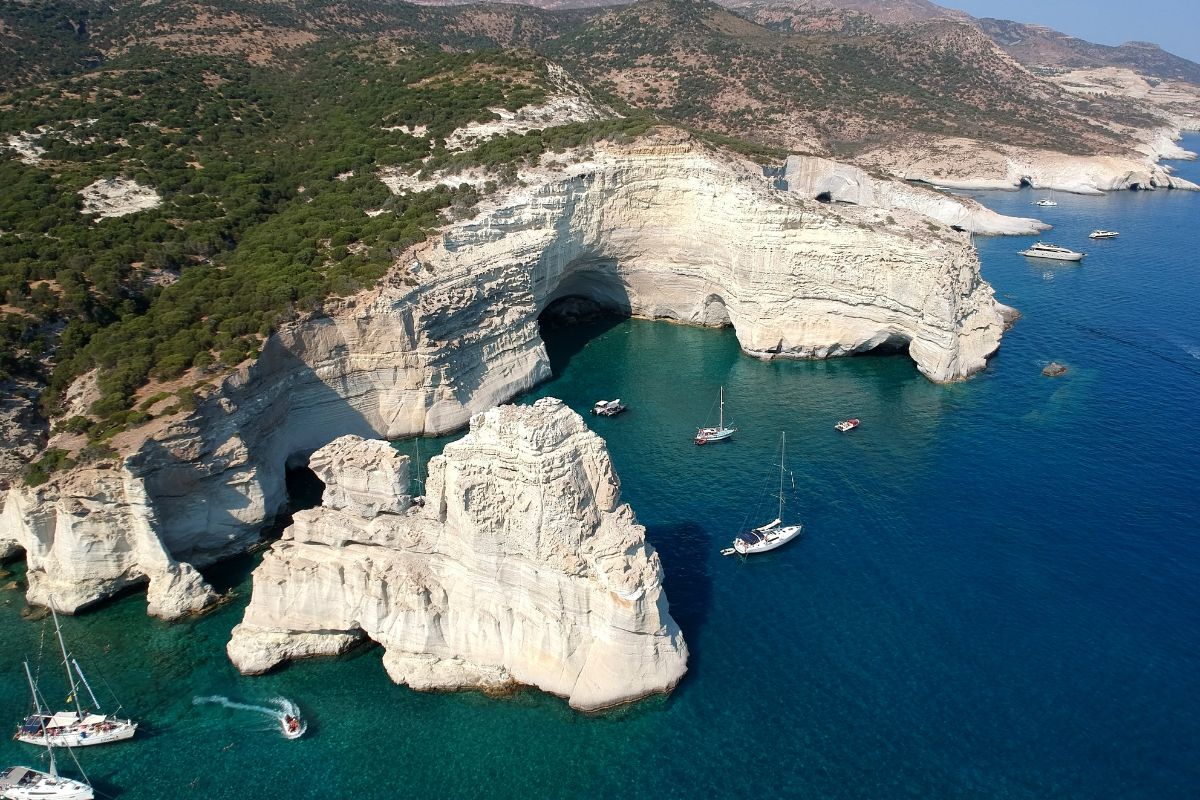 A stunning view of Kleftiko Beach on Milos, featuring dramatic white rock formations rising from the clear turquoise water, with several sailboats anchored nearby, surrounded by a rugged coastline.