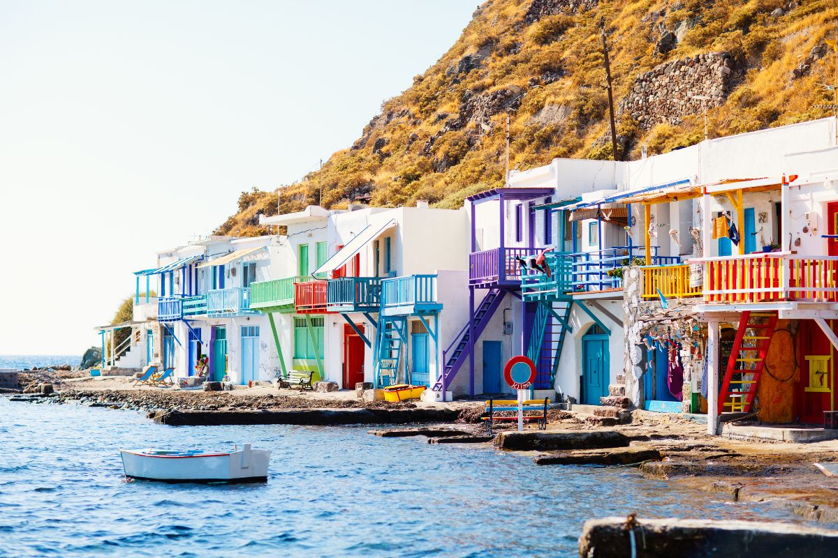 A picturesque view of Klima village on Milos, with its traditional colorful houses lining the water's edge, contrasting against a rocky hillside. A small boat floats nearby in the calm sea.