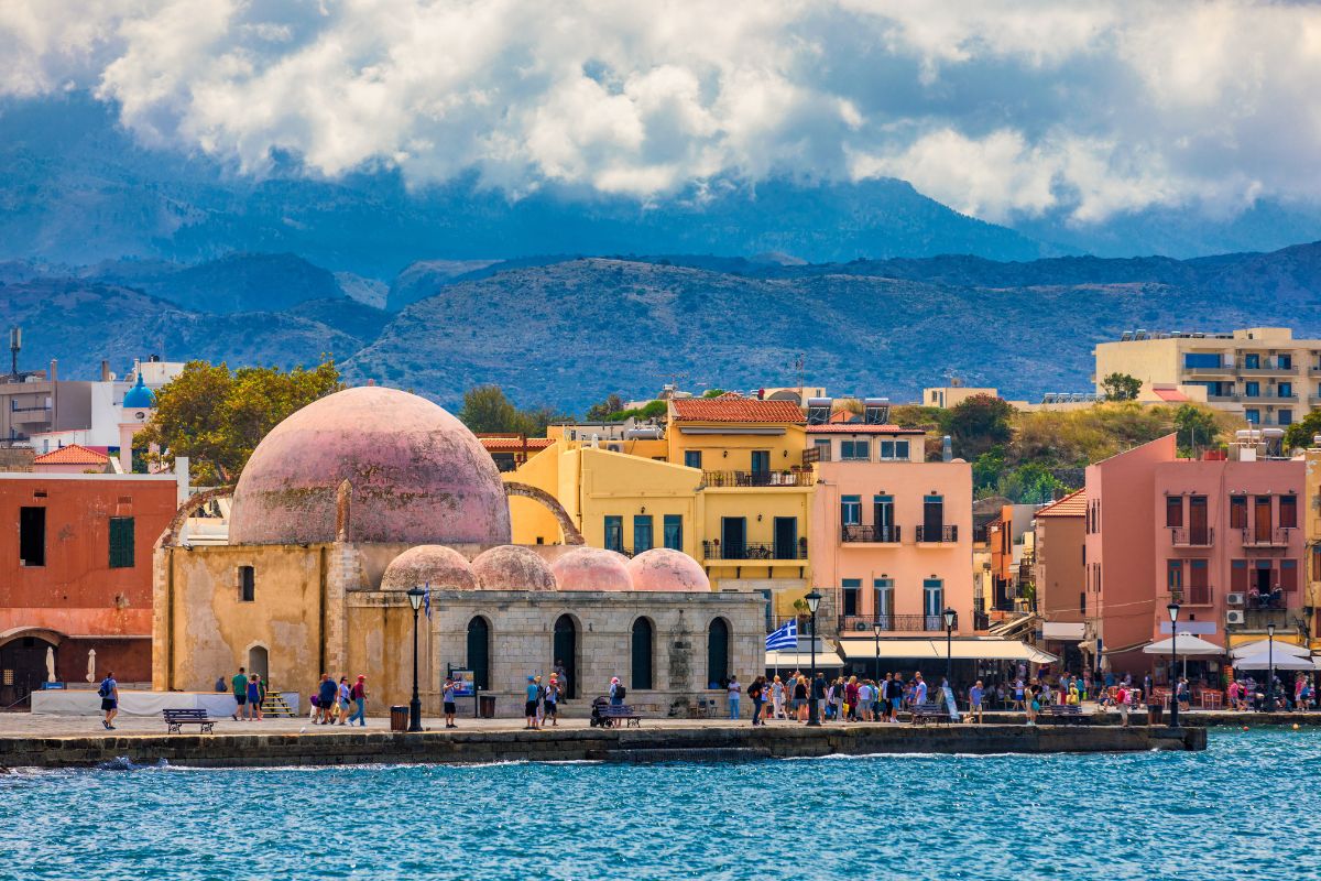 A picturesque view of Chania in winter, showcasing the iconic mosque, colorful waterfront buildings, and dramatic mountain scenery under a cloudy sky.