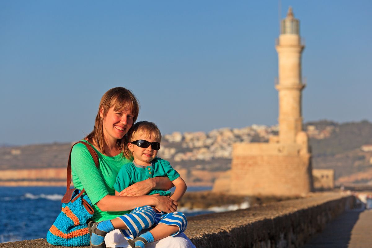 A smiling woman and child in matching green outfits sit by the waterfront in Chania, Crete, with a historic lighthouse and hillside buildings in the background.