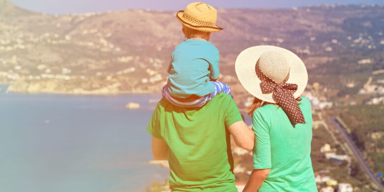 A family wearing summer hats and green shirts enjoys a scenic view of the coastline and mountains in Crete, with a child sitting on a parent's shoulders.