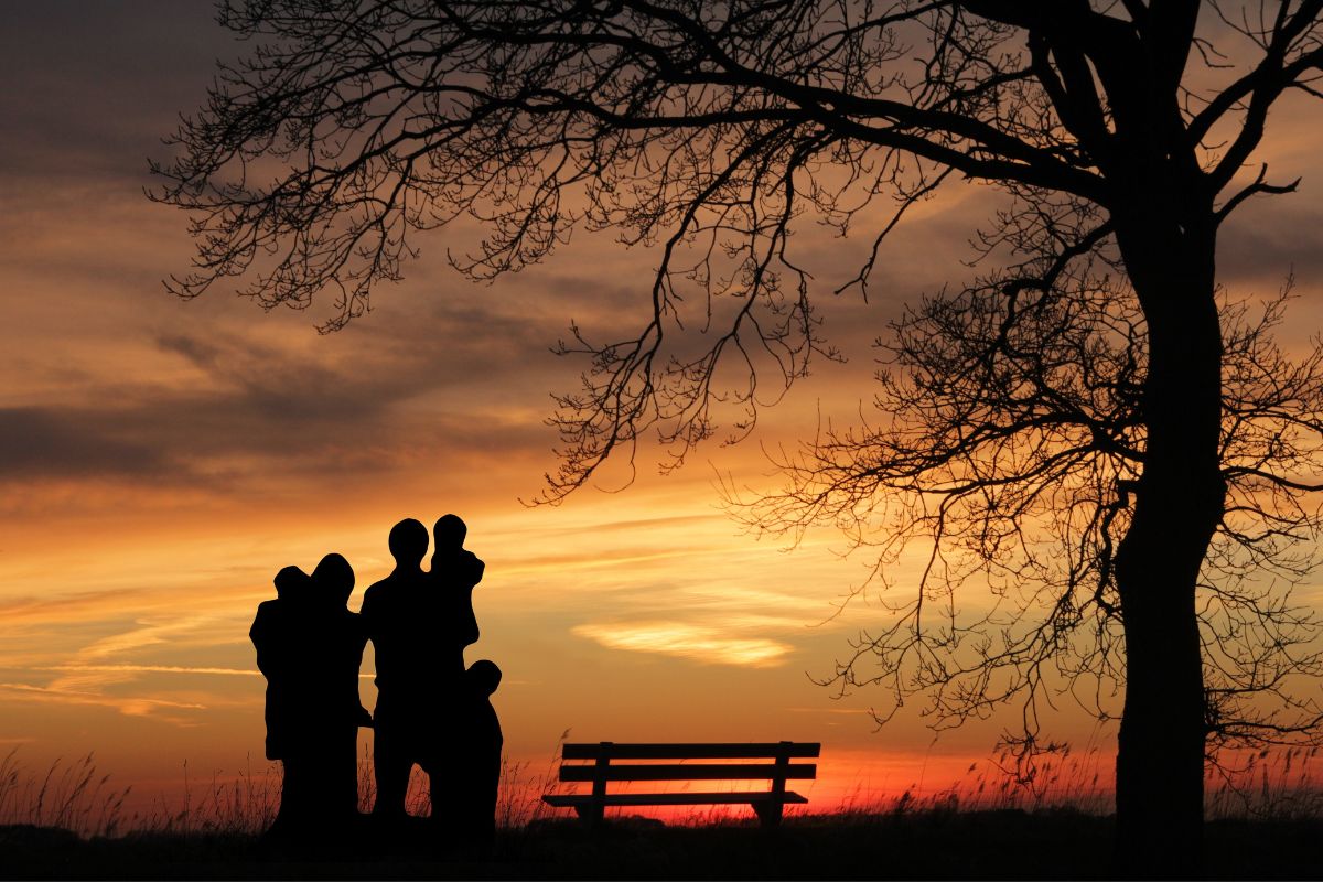 A silhouetted family stands near a bench under a tree, admiring a vibrant sunset in Crete.