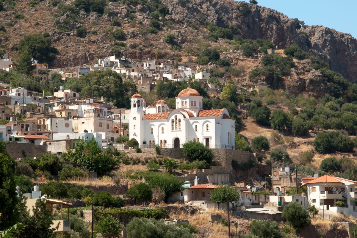 A picturesque image of Kritsa Village in Crete nestled in a hillside, featuring a striking white church with orange domes surrounded by rustic houses and lush greenery.