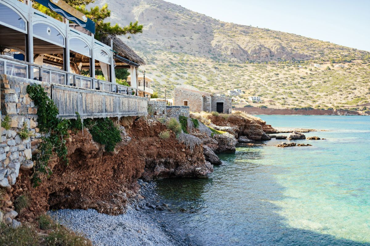 A seaside restaurant in Plaka Crete perched on rocky terrain with crystal-clear water below and hills in the background.