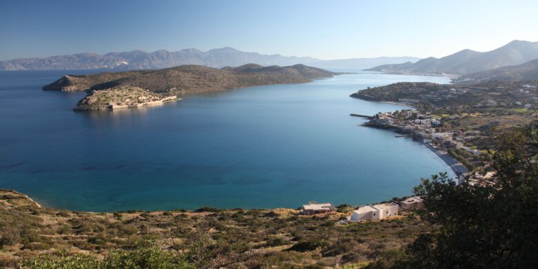 A panoramic view of a serene coastal landscape with small islands, a quiet village, and mountains in the distance. plaka crete