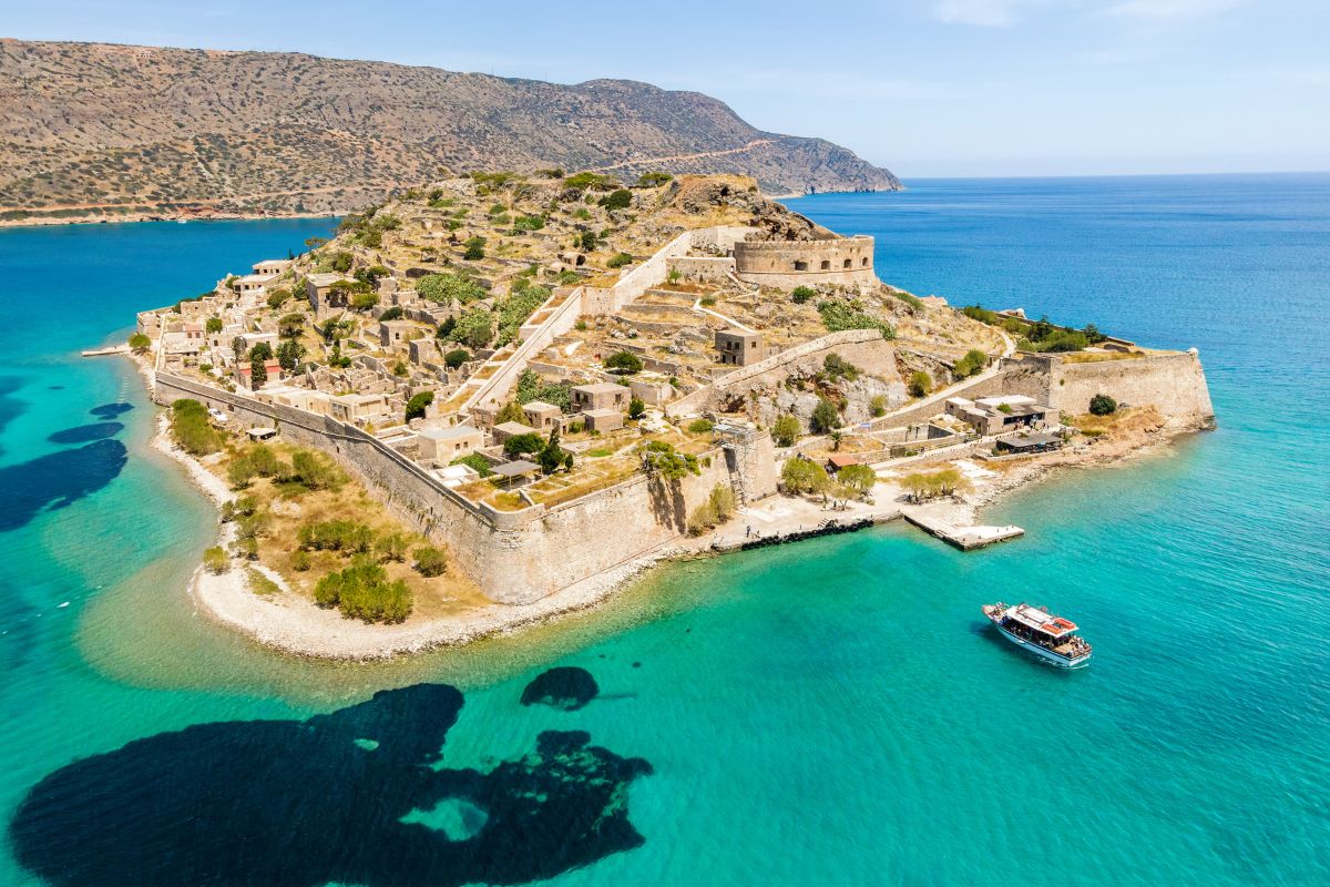 An aerial view of a historic island fortress spinalonga island surrounded by turquoise waters, with a small boat nearby.